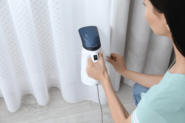 Woman using a handheld steamer to iron washable curtains near a window in a cozy Dubai home, 2025 modern interior.