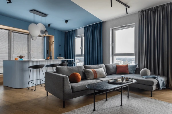 Modern Living Room with Blue & Grey Curtains in Dubai 2025, featuring a cozy grey sofa, colorful cushions, and elegant floor-length drapes complementing a chic open kitchen.