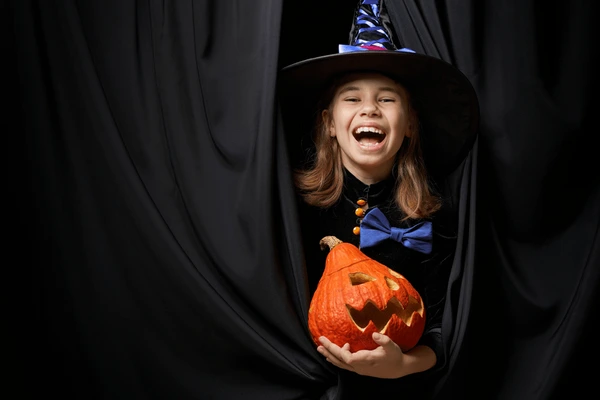 Smiling girl in a witch costume holding a carved pumpkin in front of elegant black Halloween curtains in Dubai.